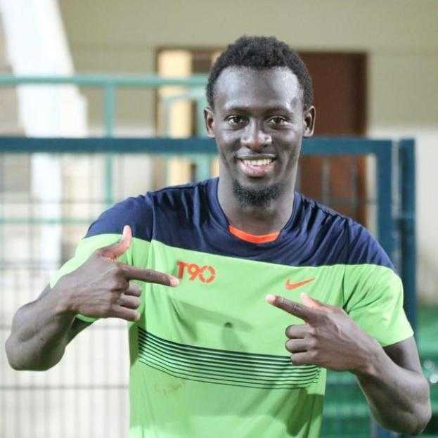 Ein Mann in einem grünen und blauen Hemd, der fröhlich in die Kamera zeigt und auf eine unscharfe Brüstung, Wand und Tür im Hintergrund zeigt.