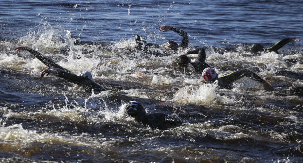 Gruppe von Menschen, die in Formation schwimmen, während eines Triathlons, tragen schwarze Neoprenanzüge und Helme, mit reflektierendem Sonnenlicht auf dem Wasser.