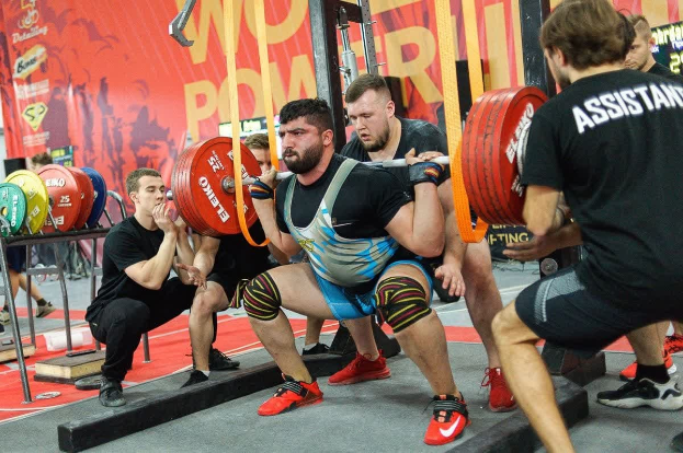 Gruppe von Männern führt Barbell-Squats in einem Fitnessstudio während der Powerlifting-Weltmeisterschaften 2019 durch, umgeben von Fitnessgeräten und Bannern.
