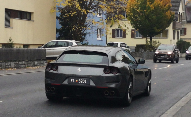 Roter Ferrari GTC4Lusso auf einer Straße mit Gebäuden, Bäumen und einem Zaun im Hintergrund unter einem sichtbaren Himmel.
