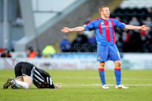 Ein Stadion mit einer Person, die steht, und einer anderen, die auf dem Rasen kniet, sowie einer Gruppe von Menschen mit Schildern im Hintergrund.