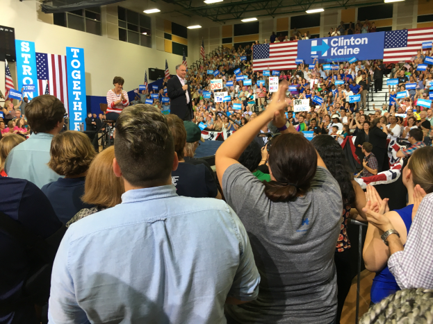 Eine große Menschenmenge steht vor einer Bühne, auf der ein Mann in ein Mikrofon spricht und eine Frau neben ihm steht, während Fahnen, Banner und ein Lautsprecher auf der Rückseite der Bühne zu sehen sind und die Menge klatscht und jubelt.