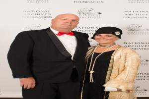 Ein Mann in einem schwarzen Anzug mit einem weissen Hemd und einer schwarzen Fliege steht neben einer Frau in einem eleganten Kleid mit einem gemusterten Schal auf einem roten Teppich, mit einem weissen Banner mit Text im Hintergrund.