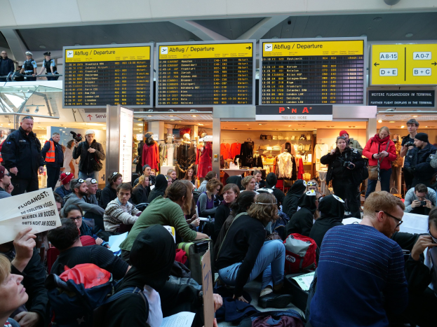 Große Gruppe von Menschen an einem Flughafen, einige mit Taschen und Papieren sitzend, andere stehend, mit Texttafeln, Schaufensterpuppen in Kleidern und Deckenbeleuchtung im Hintergrund, was auf eine Demonstration hindeutet.