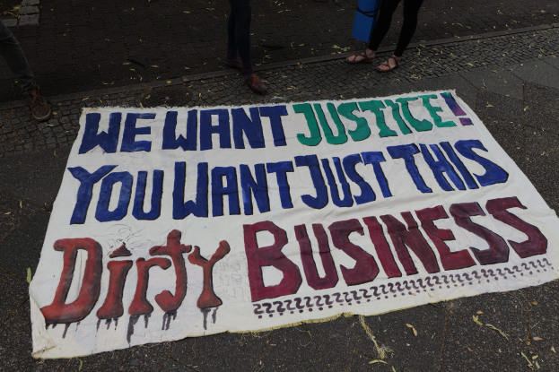 Banner auf einer Straße mit dem Text "Wir wollen Gerechtigkeit, du willst nur diesen schmutzigen Deal" und zwei Personen, eine hält ein blaues Objekt.