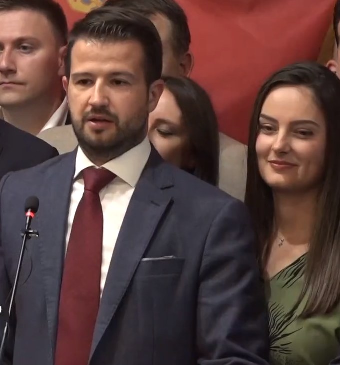 Ein Mann in Anzug und Krawatte steht an einem Rednerpult mit einem Mikrofon, spricht zu einer Menge mit einem Plakat im Hintergrund, wahrscheinlich auf eine Regierungssperre für soziale Medien reagierend.