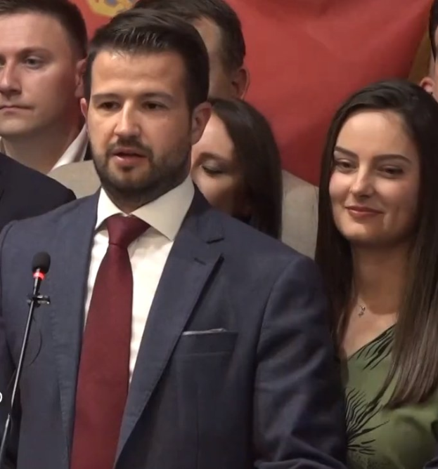 Ein Mann in Anzug und Krawatte steht an einem Rednerpult mit einem Mikrofon, spricht zu einer Menge mit einem Plakat im Hintergrund, wahrscheinlich auf eine Regierungssperre für soziale Medien reagierend.