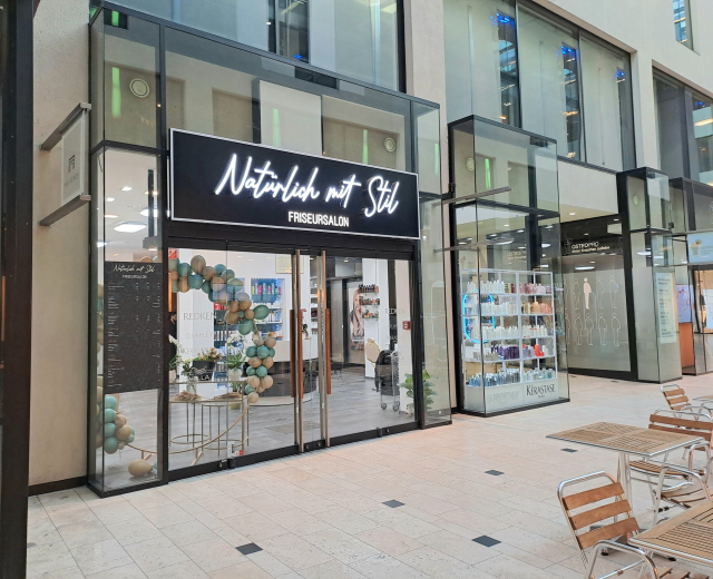 Storefront of Naturlich mit Stil hair salon with outdoor seating, glass walls, and a signboard; inside, shelves display bottles and other items.