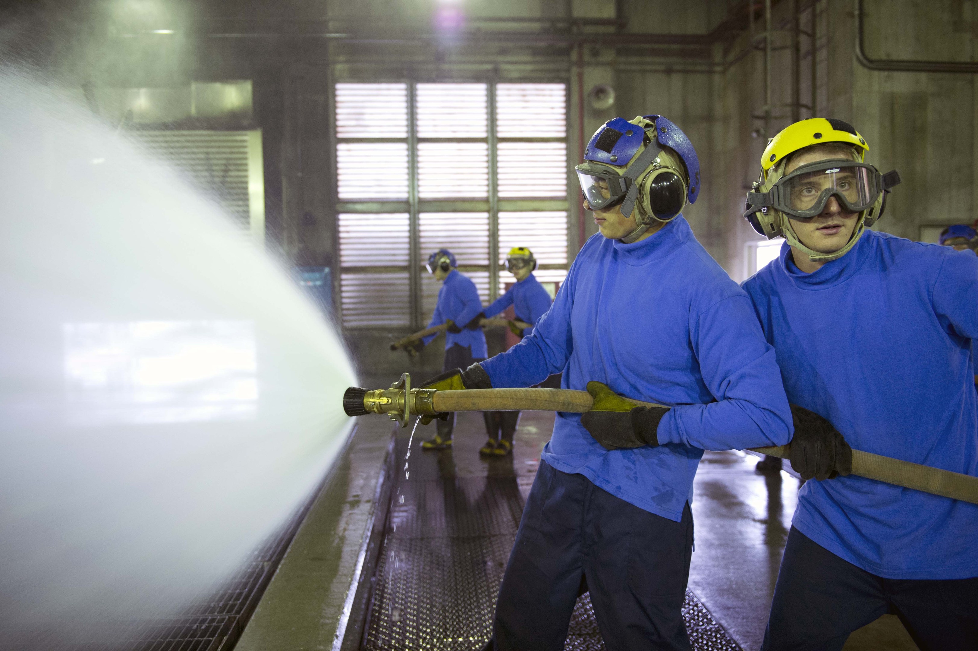 Männer in blauen Hemden und gelben Helmen arbeiten an Maschinen, einer sprüht Wasser auf den Boden, in einer Fabrikumgebung mit Wänden, Fenstern, Rohren und Lampen.