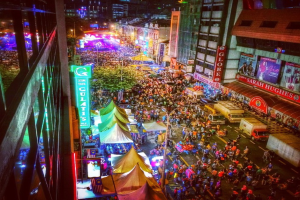 Ein buntes Karnevalszenario mit einer großen Menge auf der Straße, umgeben von Zelten, Geschäften, Gebäuden und Bannern, mit zahlreichen Lichtern im Hintergrund.