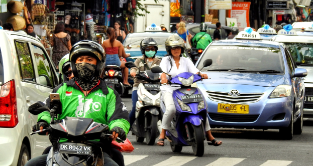 Eine Gruppe von Menschen auf Motorrädern auf einer vielbefahrenen Straße mit Autos, Gebäuden, Bäumen und Geschäften im Hintergrund.