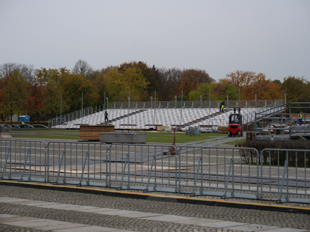 Großes Stadion mit Metallgeländern, umgeben von Bäumen unter einem klaren blauen Himmel, mit Menschen und Fahrzeugen im Vordergrund auf saftig grünem Gras.