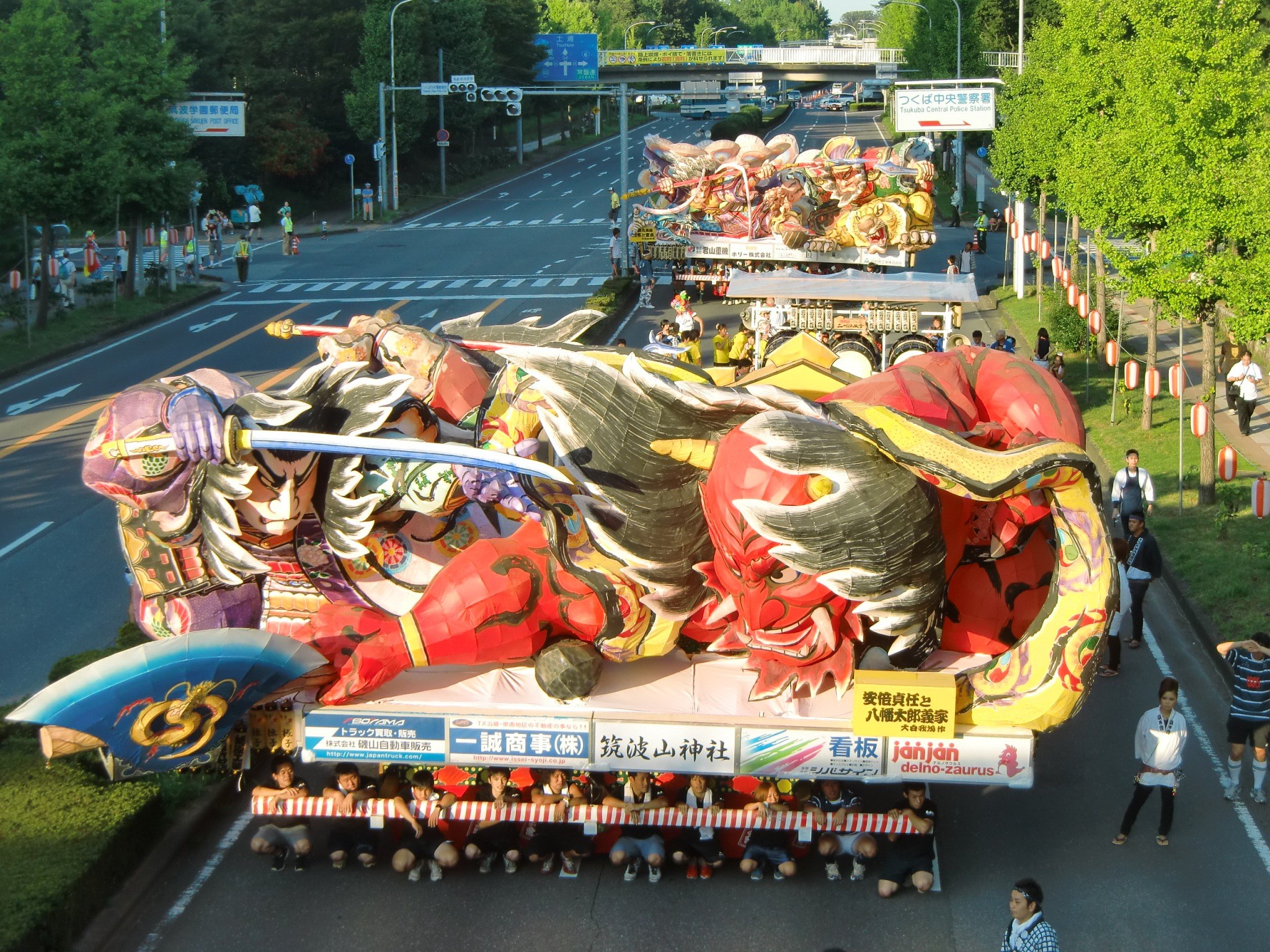 Eine Gruppe von Menschen auf einem geschmückten Festwagen in einem Umzug, umgeben von Bäumen, Laternenmasten und einer Brücke, mit ein paar Zuschauern rechts daneben.