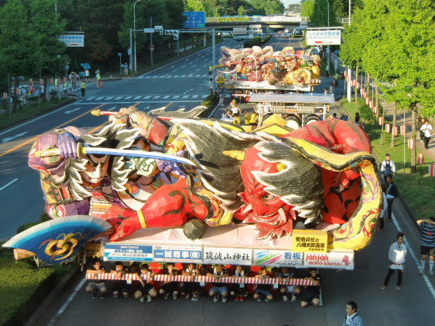 Eine Gruppe von Menschen auf einem geschmückten Festwagen in einem Umzug, umgeben von Bäumen, Laternenmasten und einer Brücke, mit ein paar Zuschauern rechts daneben.