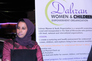 Eine Frau in einem weißen Hemd und blauen Jeans steht entschlossen vor einer Tafel mit der Aufschrift "Dahran Frauen & Kinderbefähigung", mit einem Auto im Hintergrund.