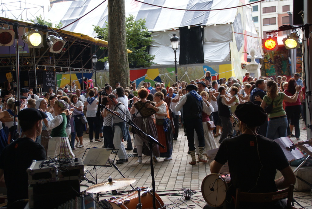 Eine Gruppe von Menschen tanzt im Freien mit einem Mann, der rechts ein Drum spielt, einem Mann, einem Licht, einem Baum und einem Zelt links und einem Gebäude mit einer Straßenlaterne im Hintergrund.