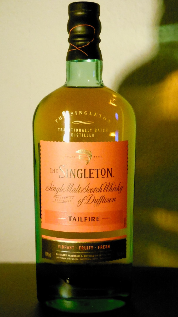 Eine tiefe bernsteinfarbene Flasche Singleton of Dufftown Tailfire Single Malt Scotch Whisky auf einem Tisch, mit einem Etikett, das eine schwarze Schrift, ein Löwenwappen und eine Burg im Hintergrund zeigt.