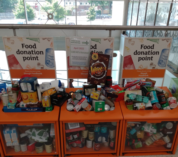 Orange Schübe mit Lebensmitteln wie Flaschen, Dosen und Kisten an einer Spendenstelle, mit Informationsschildern in der Nähe und einem Blick durch Glasfenster auf Außenfahrzeuge, Bäume, Gebäude und Himmel.