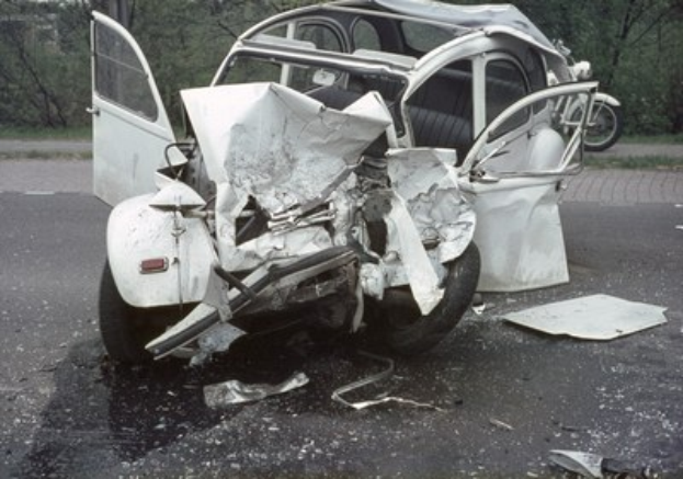 Ein schwerbeschädigtes Auto mit eingedrückter Front steht am Straßenrand, mit Bäumen und einem Gebäude im Hintergrund und einem weiteren Fahrzeug in der Ferne sichtbar.