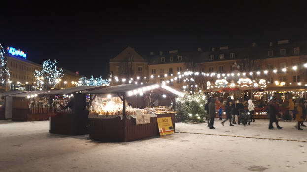 Ein nächtlicher Weihnachtsmarkt auf einer verschneiten Straße mit Menschen, Buden, Pflanzen, Bäumen, Gebäuden und Schildern unter einem bewölkten Himmel.
