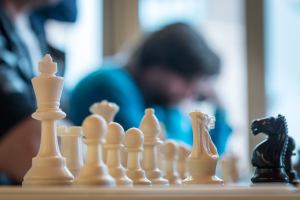 Ein Schachbrett mit Figuren und ein paar Personen im Hintergrund.