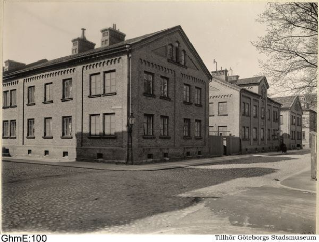 Black and white photograph of a street with buildings, street poles, street lights, vehicles, trees, and sky, with "Tillhör Göteborgs Stads Museum" at the bottom.