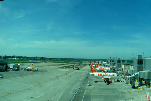 Eine Flughafen-Szene mit Flugzeugen, Gebäuden, Polen, Fahrzeugen und Bäumen unter einem Himmel mit einer Straße unten und einem Zaun im Vordergrund.