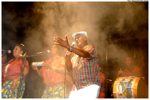 Eine Gruppe von Musikern, die auf der Bühne spielen, mit einem Mann, der in ein Mikrofon singt, in der Mitte und einem anderen Mann, der rechts ein Schlagzeug spielt.