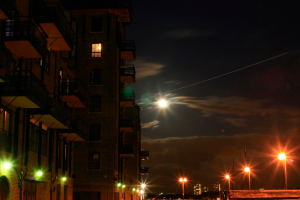 Eine nächtliche Stadtansicht mit einem prominenten Gebäude mit zahlreichen Fenstern und Balkonen, wobei auf der rechten Seite Straßenlaternen zu sehen sind.