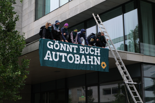 Eine Gruppe von Menschen auf einem Gebäude mit einem Banner, mit einem Baum links und Glasfenstern im Hintergrund, wahrscheinlich protestierend für die deutsche Regierung.