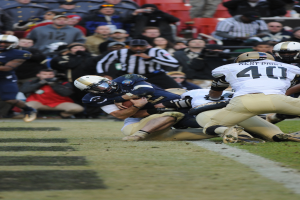 Ein Fussballspieler wird von einer Gruppe von Verteidigern auf einem grünen Feld getackelt, mit Zuschauern und Kameramännern im Hintergrund, alle Spieler tragen Helme.