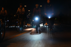 Eine Gruppe von Menschen, die nachts auf Fahrrädern durch eine Stadtstraße fahren, mit Helmen und beleuchtet von Stadtlichtern.