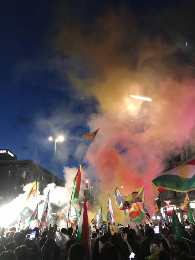 Eine Menschenmenge vor einem Gebäude, die Fahnen schwenkt und Handys hält, mit farbigen Feuerwerken am Himmel.