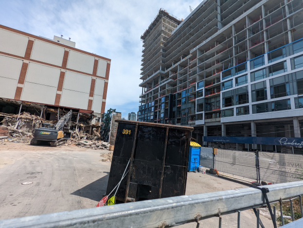 Großes Gebäude mit erheblichen Trümmern davor, umgeben von einem Metallzaun, umgeben von einem Container, Pflanzen, Bäumen, Pfählen, einem Schild, Fahrzeugen, Steinen und einem bewölkten Himmel, was auf eine Baustelle für einen neuen Wohnkomplex hinweist.