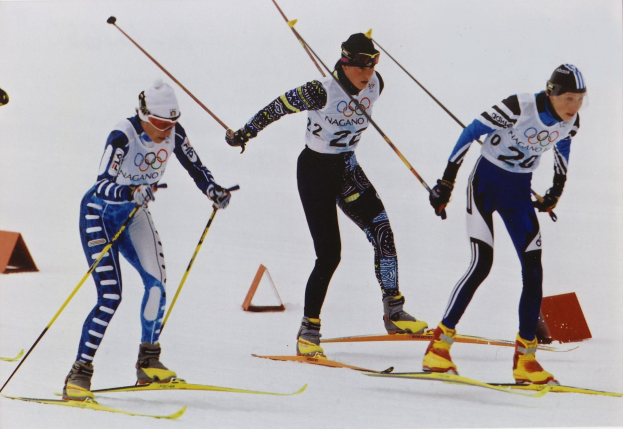 Drei Skifahrer in bunten Anzügen und Helmen rasen eine glitzernde schneebedeckte Piste hinunter, jeder hält Ski-Stöcke in der Hand.