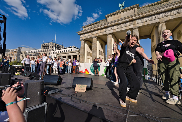 A group of people on a stage with microphones and cameras in front of the Brandenburg Gate, Berlin, with equipment and buildings in the background.