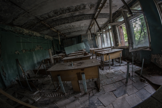Verlassener Klassenraum in Pripyat, Tschernobyl, Ukraine, mit Tischen, Stühlen, abblätternder Wandfarbe, kaputten Fenstern und sichtbarer Decke, Bäume im Hintergrund durch Fenster zu sehen.