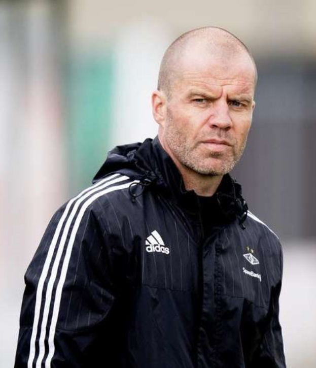 Ein kahlköpfiger Trainer in einer blauen Jacke mit weißen Streifen und einem weißen Kragen steht auf einem Fußballfeld und schaut ernsthaft nach vorne.