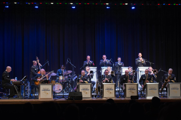 Eine Gruppe von Musikern performs auf einer hell erleuchteten Bühne mit Mikrofonen und Ständern, begleitet von Texttafeln mit der Aufschrift "The New Orleans Jazz Band" und einem sichtbaren Publikum darunter.