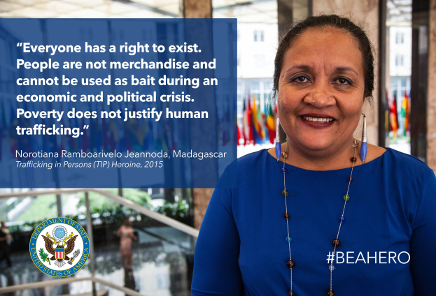 Eine Frau in einem blauen Kleid steht lachend vor einem Gebäude mit einer Inschrift über den Menschenhandel, umgeben von Menschen, Pflanzen und Flaggen.