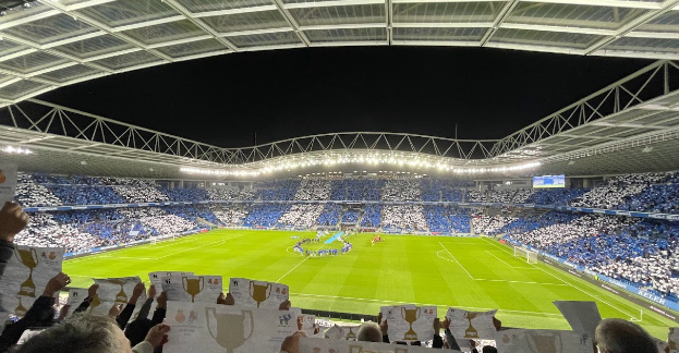 A group of people holding signs in front of a soccer stadium, with a few standing in the foreground and others seated in the background under stadium lights, suggesting a celebratory victory event.
