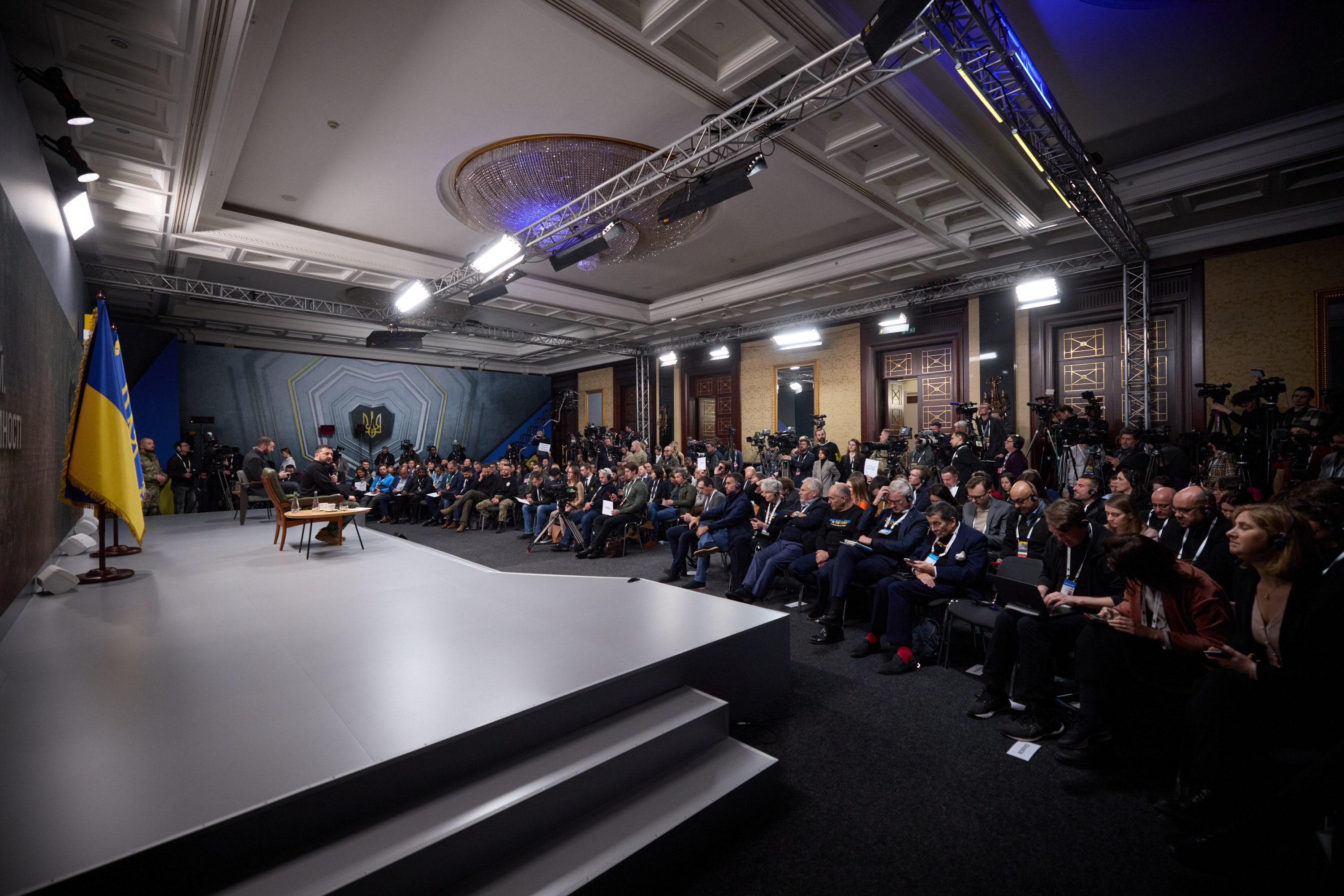 Große Gruppe von Menschen auf Stühlen sitzend, die einer Bühne mit zentralem Tisch, einer Fahne links und Fotografen rechts zugewandt sind, in einem Raum mit Deckenleuchten und Türen.