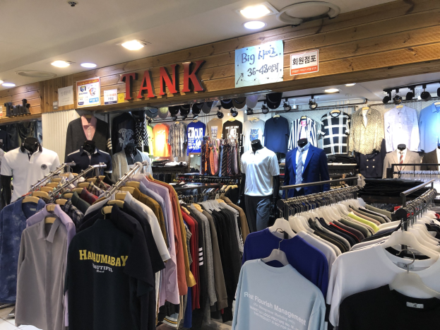 Ein modernes Bekleidungsgeschäft mit zahlreichen auf Ständern präsentierten Shirts, Schaufensterpuppen, die aktuelle Outfits präsentieren, informativen Tafeln an den Wänden und Deckenbeleuchtung.