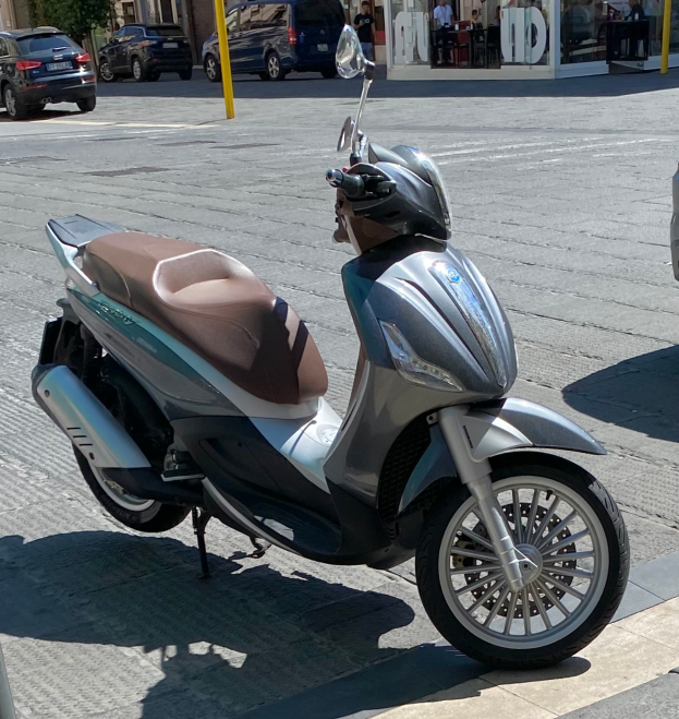 Ein Motorroller am Straßenrand mit einem Helm darauf, mit Gebäuden, Bäumen, Polen, Menschen, Stühlen, Tischen und anderen Gegenständen im Hintergrund.