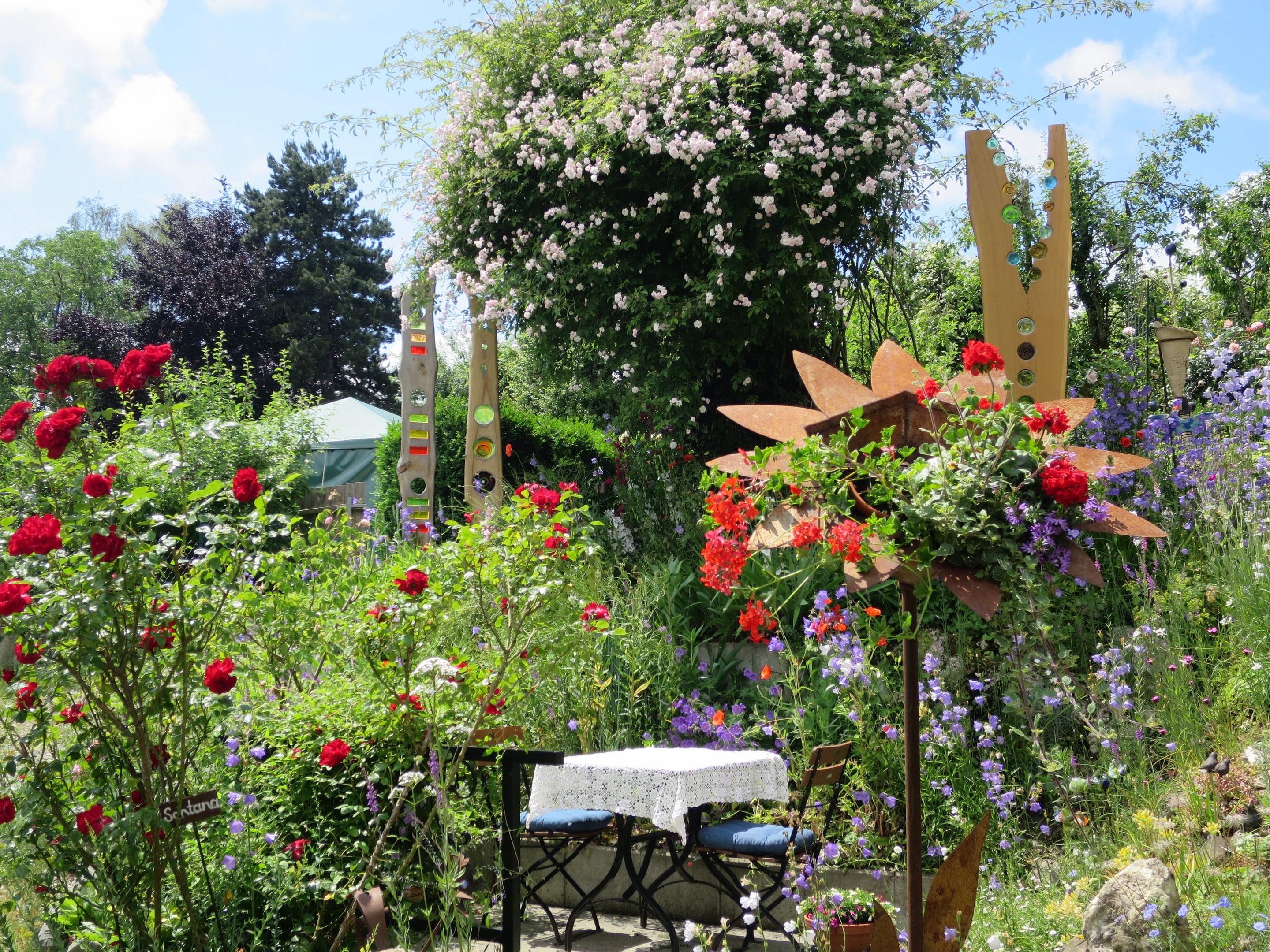 Ein Garten auf der Chelsea Flower Show mit bunten Blumen und Pflanzen, einem Tisch mit einer Decke und Stühlen in der Mitte und Bäumen gegen einen bewölkten Himmel im Hintergrund.
