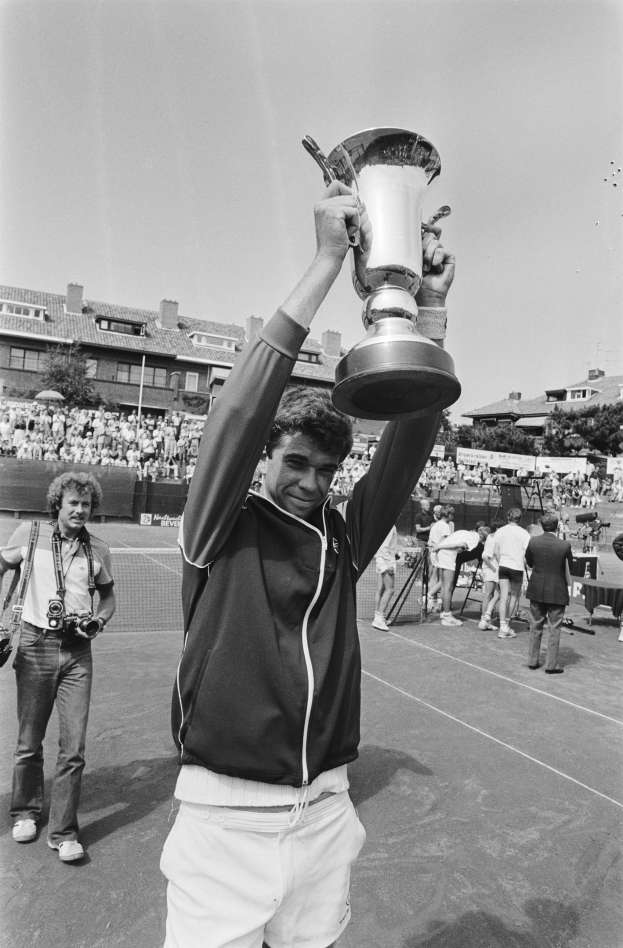 Ein Mann in Tennisbekleidung hält eine Trophäe auf einem Platz hoch, umgeben von einer jubelnden Menge mit Kameras, mit Gebäuden, Bäumen und einem klaren Himmel im Hintergrund.