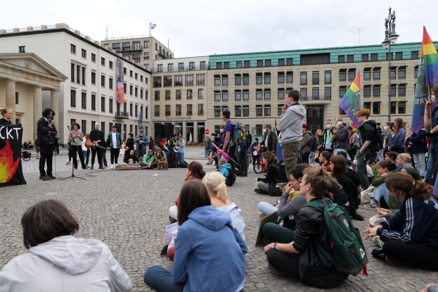 Eine Gruppe von Menschen, die auf dem Boden vor einer Menge sitzt, die Fahnen und Transparente h"alt, w"ahrend einer Anti-Schwulen-Demo in Berlin. Im Hintergrund ist eine Statue, Geb"aude und ein bew"olkter Himmel zu sehen.