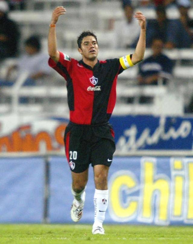 Ein Fußballspieler in rot-schwarzem Trikot rennt mit ausgebreiteten Armen auf einem Feld, umgeben von Zuschauern in den Rängen, mit einem "Chivas de Guadalajara"-Schild im Hintergrund.