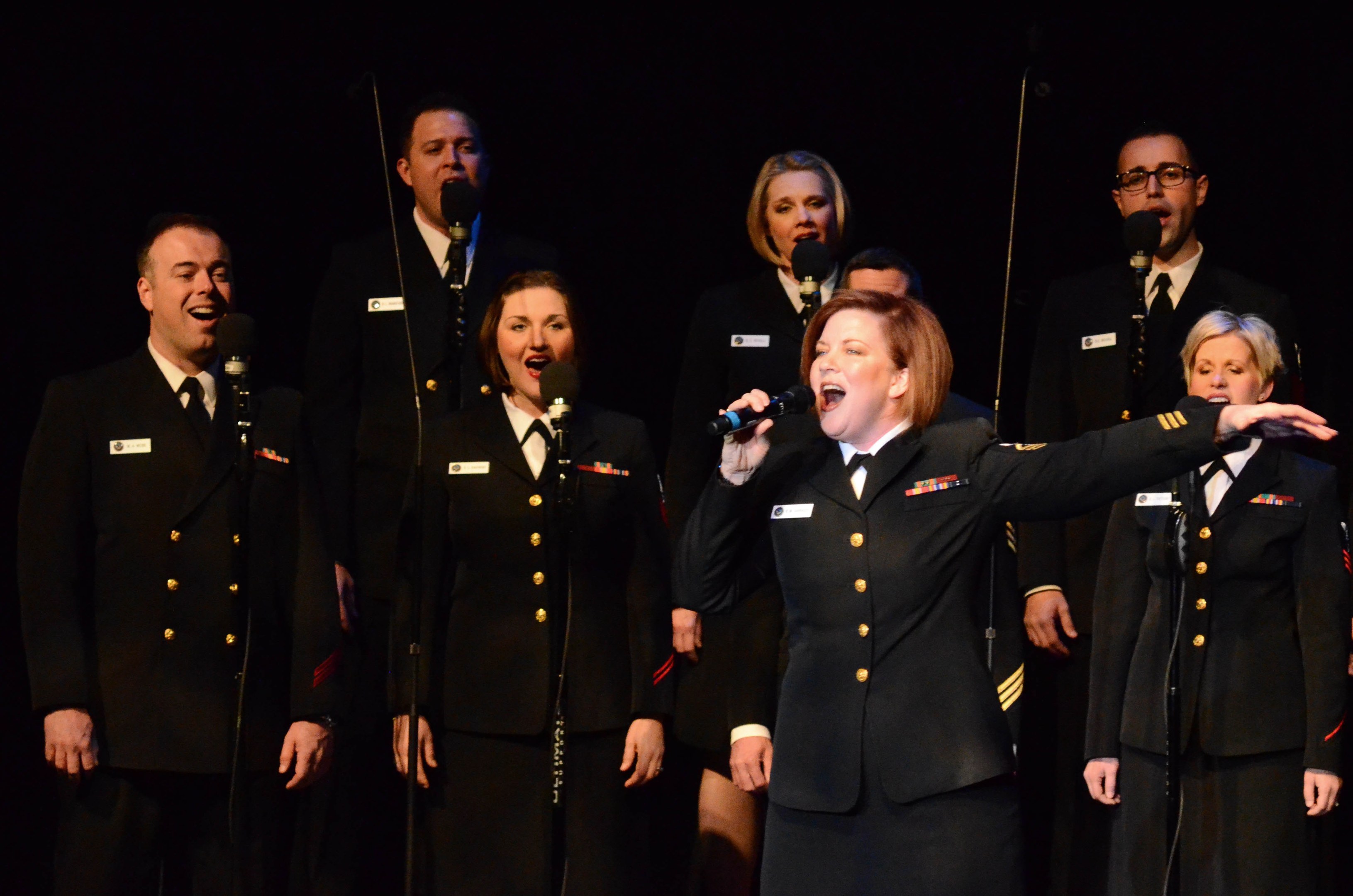 A group of people in military uniforms singing on stage with microphones, set against a dark background.