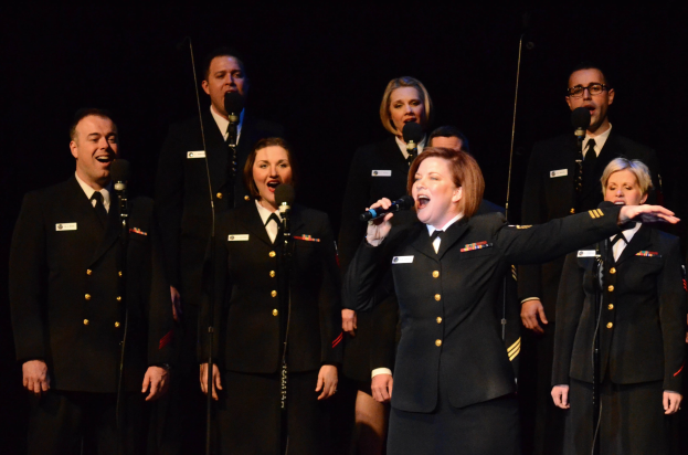 A group of people in military uniforms singing on stage with microphones, set against a dark background.